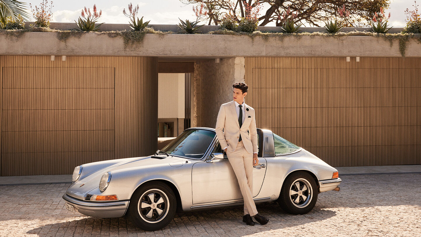 Model in POLITIX Spring Summer 2025 light beige Prince of Wales check suit with green tie, leaning on a vintage silver Porsche – modern men’s wedding and event tailoring