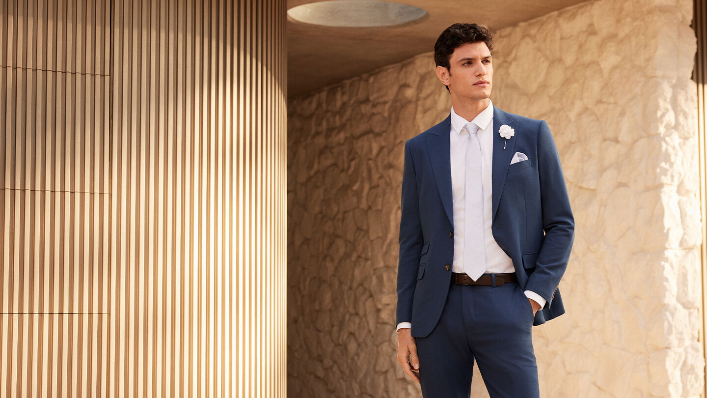Model in POLITIX Spring Summer 2025 steel blue tailored suit with white shirt, tie, and boutonnière – modern men’s wedding and eventwear.