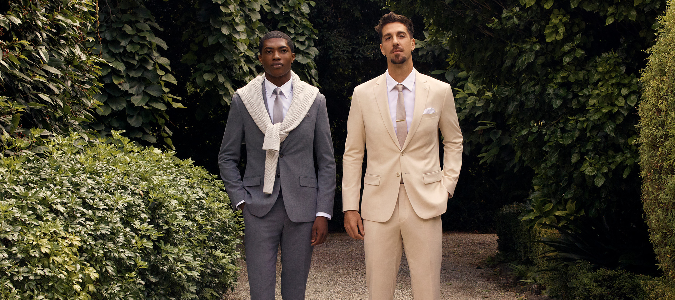 Two men wearing stylish tailored suits walking through a lush garden path. One wears a classic grey suit with a white sweater draped over his shoulders, while the other wears a light beige suit with a white shirt and tie, both styled for a refined outdoor look.