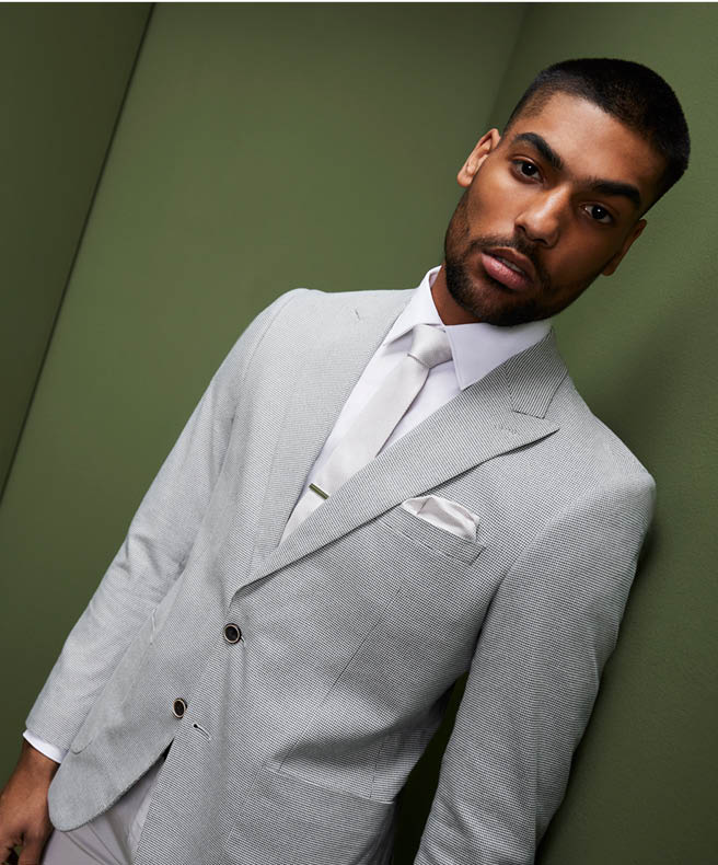 Male model wearing POLITIX light grey suit and white accessories standing in front of dark green backdrop