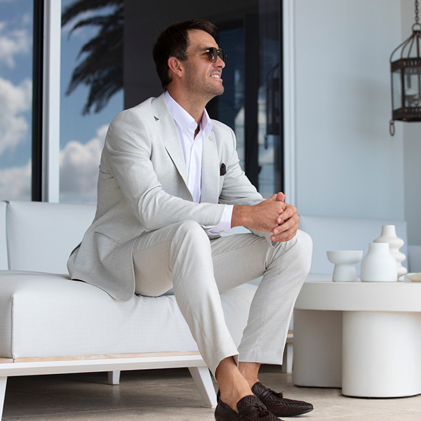 Matt Poole seated outdoors wearing cream Natural Suit Look, white shirt, brown loafers and sunglasses smiling looking away