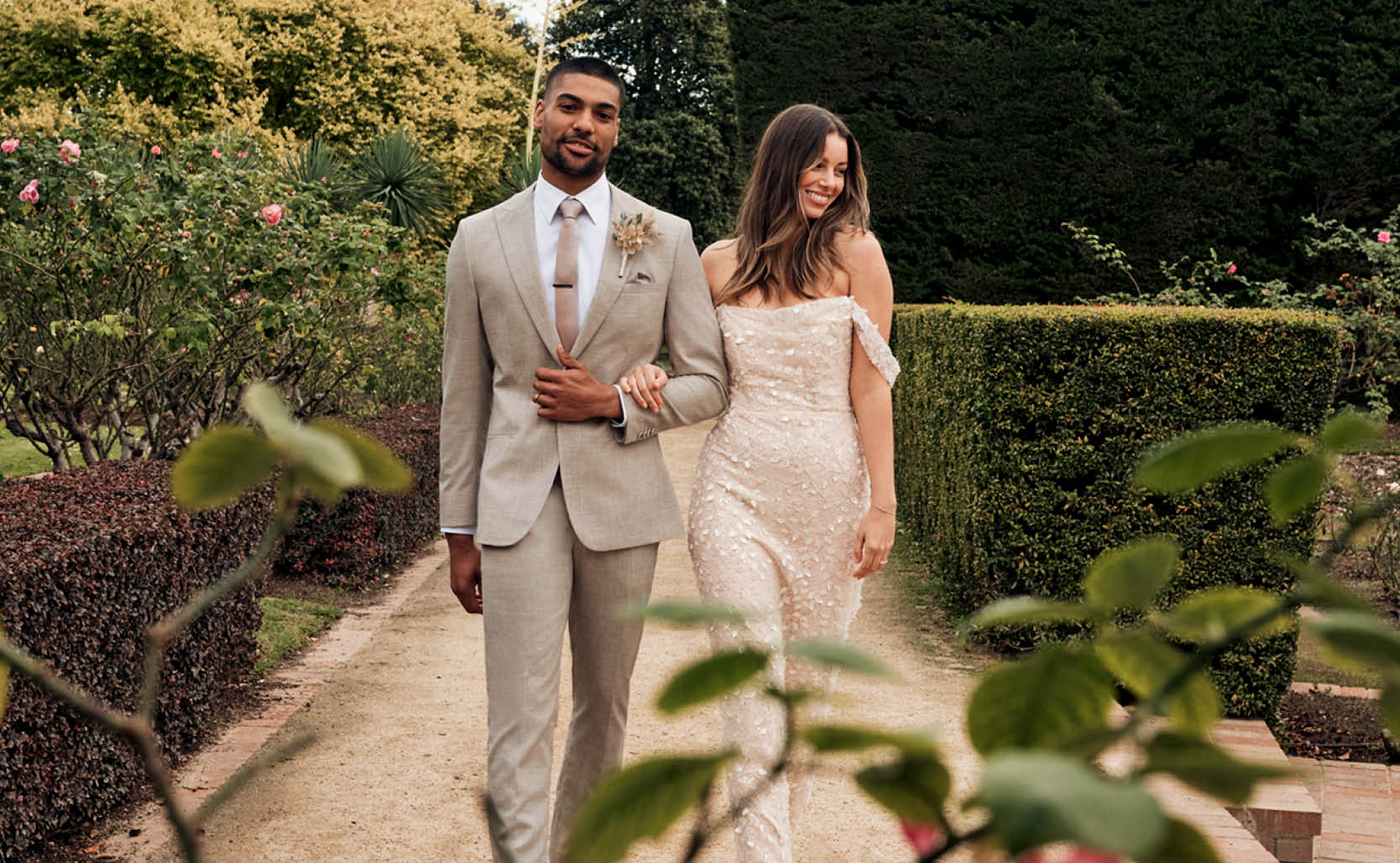 Male model wearing beige suit with female moderl wearing glittery weading dress walking through winery garden setting