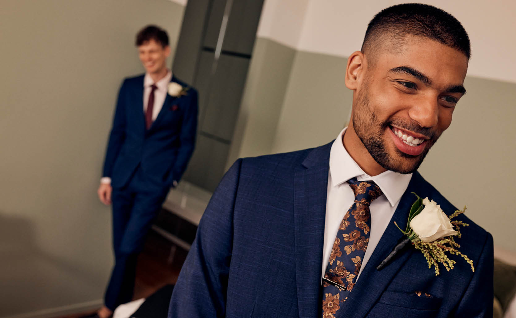 Male model smiling in foreground wearing POLITIX Navy suit with second male model in background wearing POLITIX navy blue suit