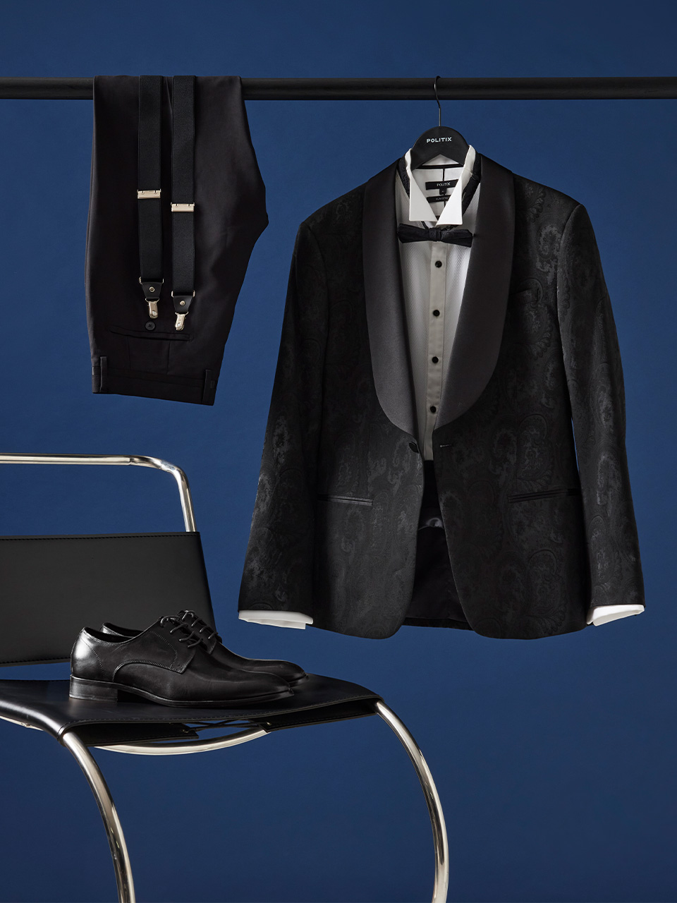 Flat lay of a black paisley tuxedo jacket with satin shawl lapel, formal trousers with suspenders, leather dress shoes, and a fly-collar shirt with bow tie from the Politix AW25 Black Tie Collection.
