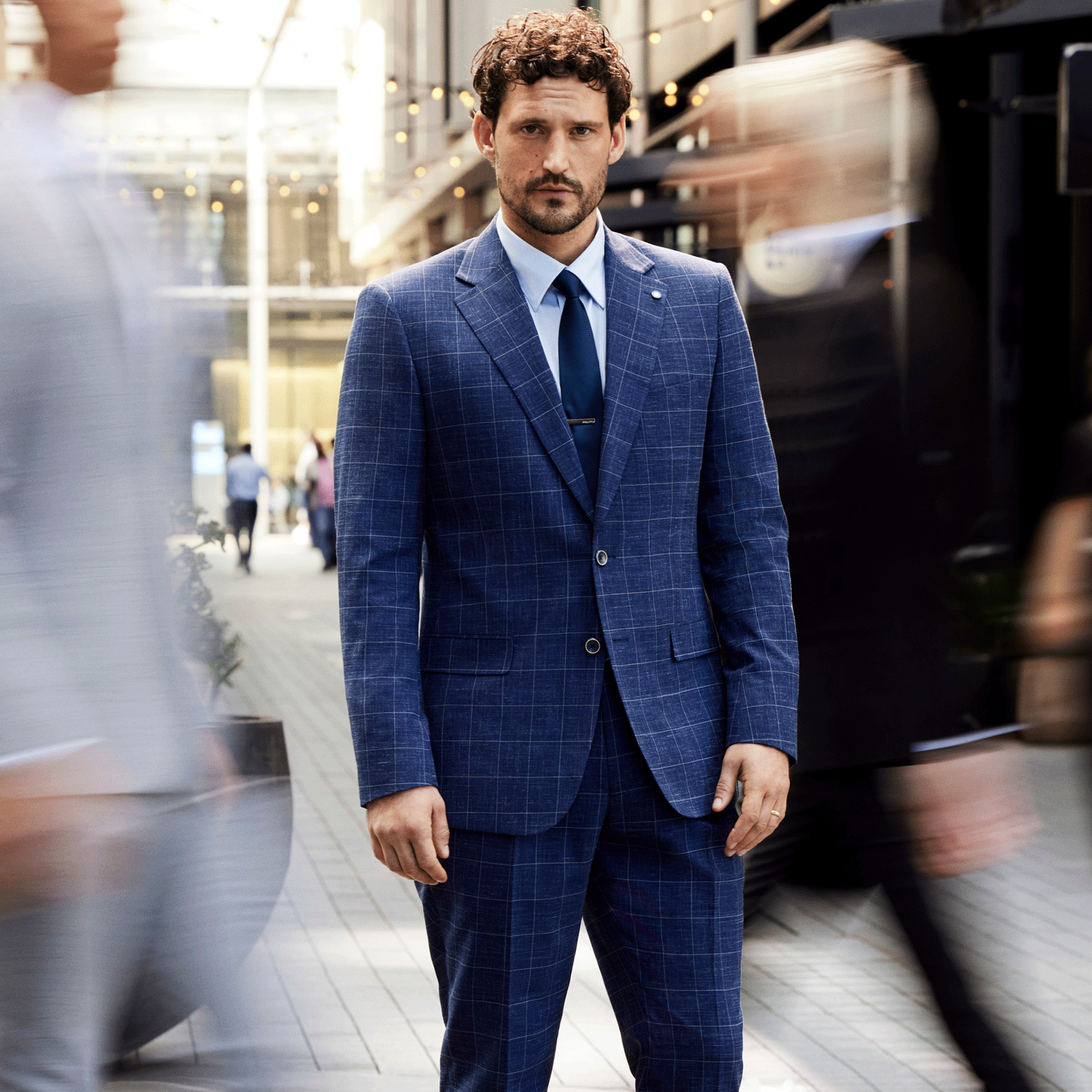 Count on a classic Model wearing POLITIX Blue Windowpane suit standing still in front of moving crowd