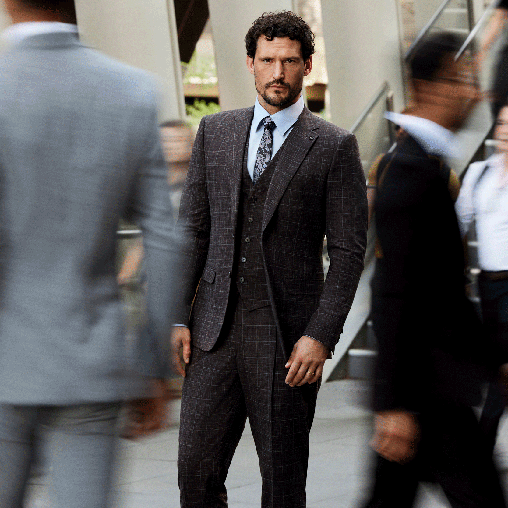 A rich tone for turning heads. Model wearing Chocolate Windowpane POLITIX suit standing in front of moving crowd