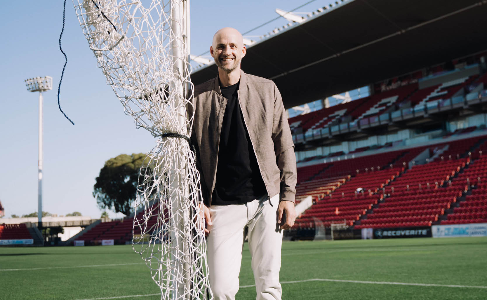 Jarrod Walsh smiling standing next to a soccer post wearing beige bomber jacket, black shirt and white chinos 