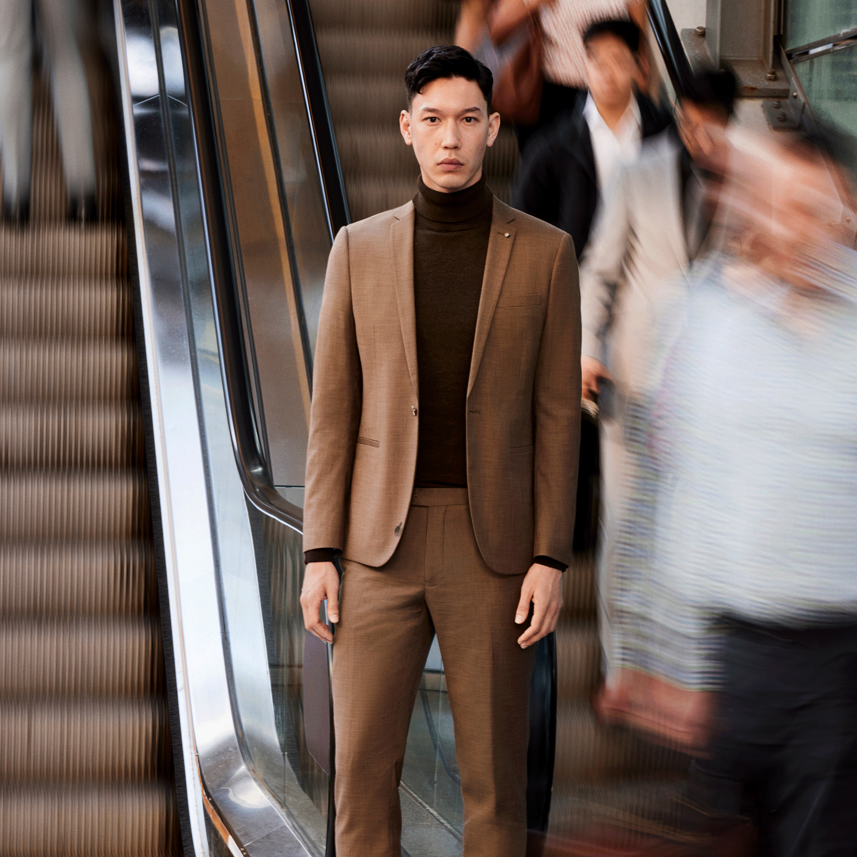 Layered to level up Model wearing Toffee POLITIX suit with chocolate roll neck standing still in front of a crowd