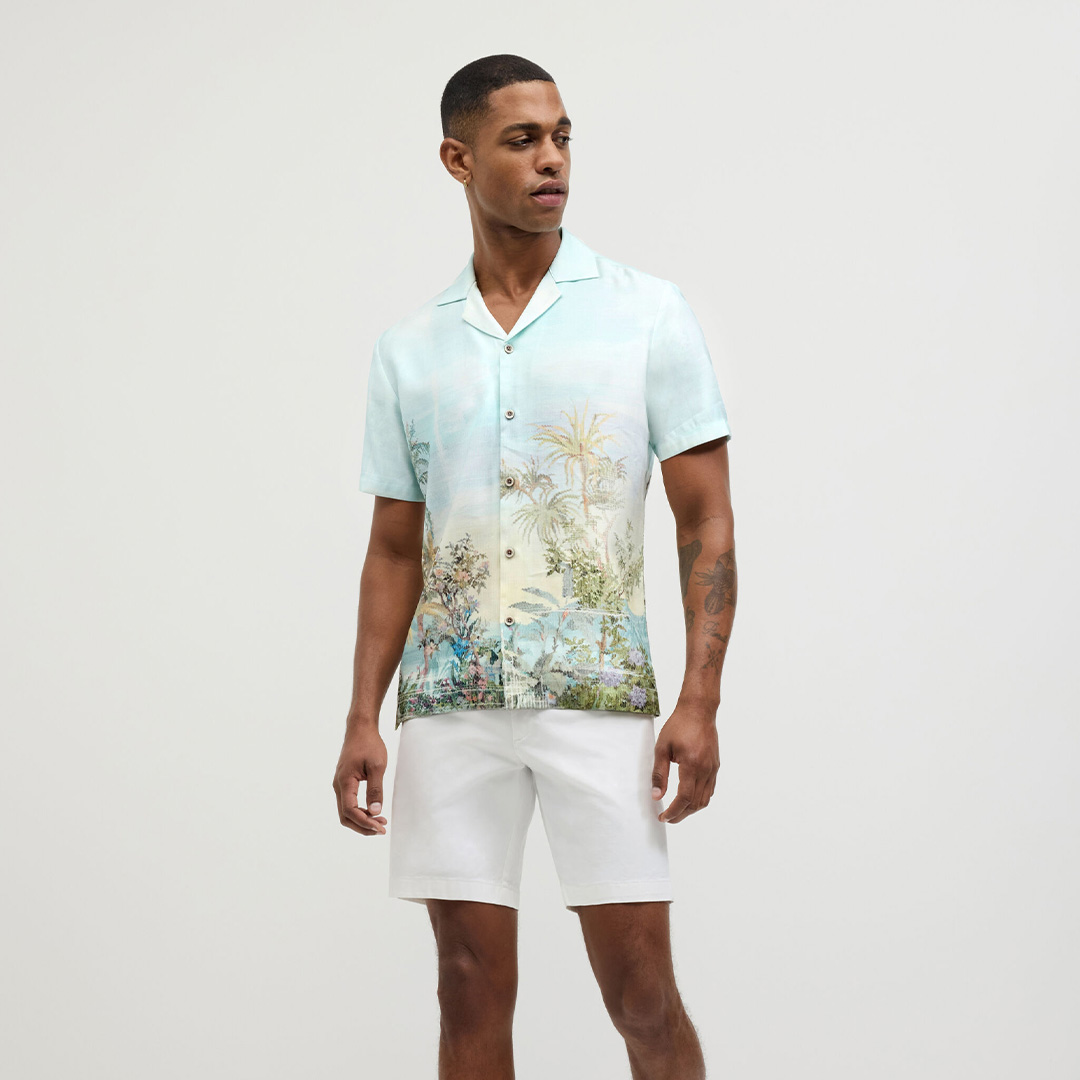 Man wearing a light-blue tropical tapestry print short sleeve shirt paired with white summer shorts.