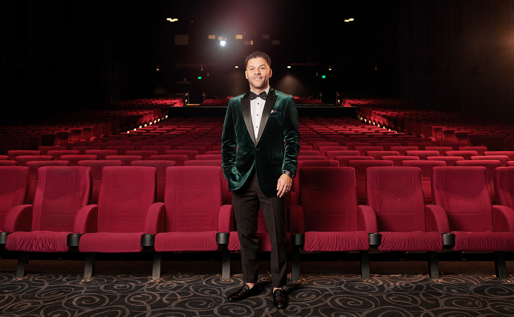 Kid Mac standing in cinema surrounded by red seats, wearing POLITIX velvet green tuxedo