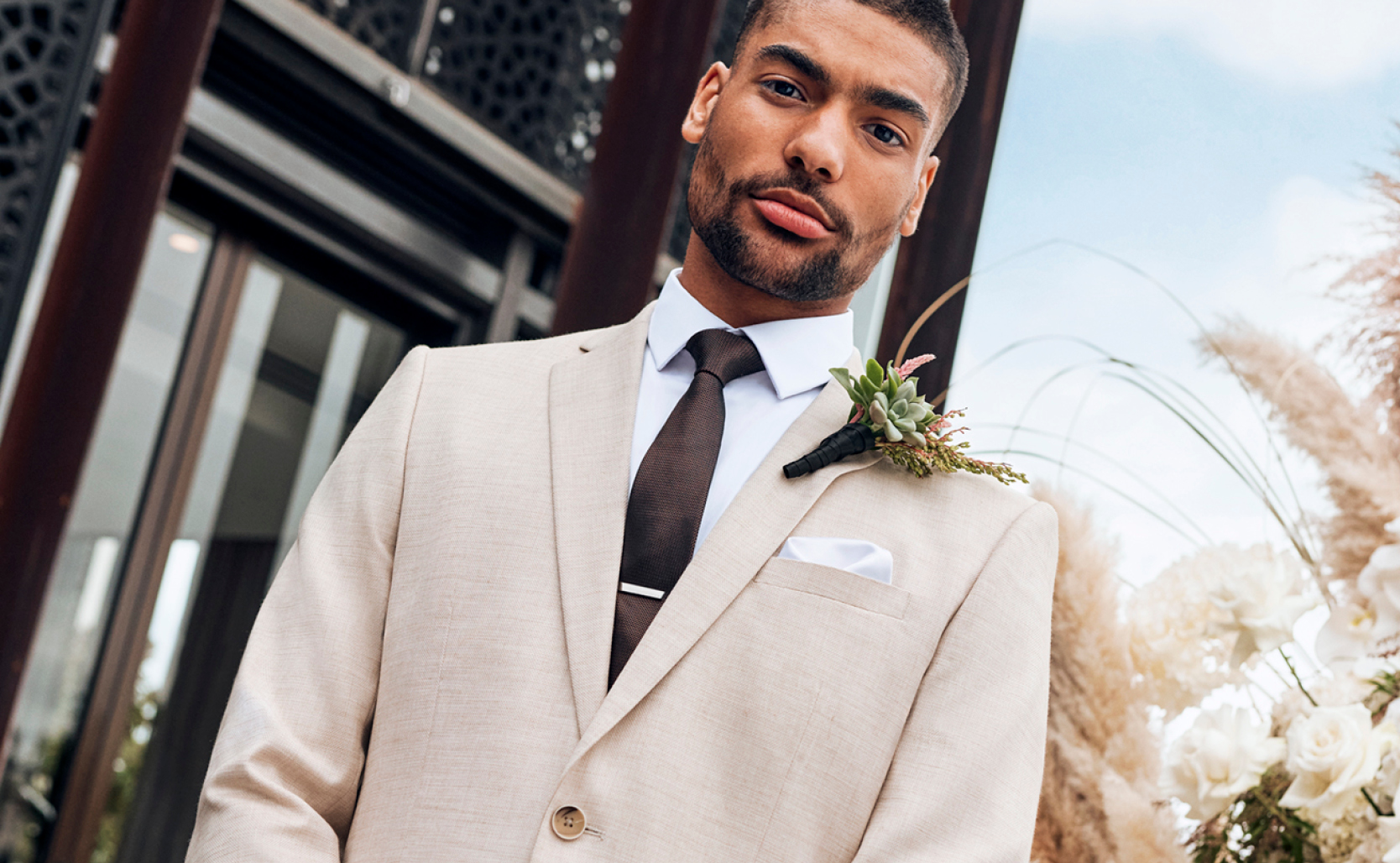 POLITIX Natural Suit Image of model on hotel deck looking into camera wearing natural coloured suit with White button up shirt and burgundy tie