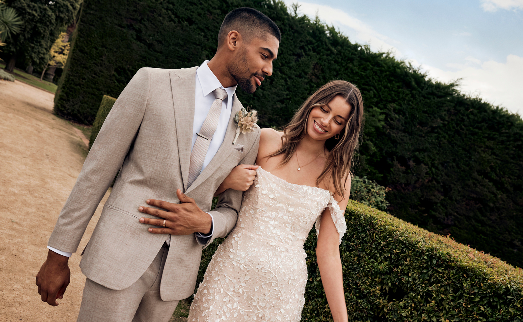 POLITIX Fawn Suit Image of male and female model walking in park. He is wearing fawn coloured suit, white button up shirt and fawn tie.