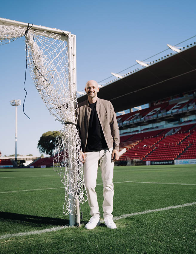 Jarrod Walsh smiling standing next to a soccer post wearing beige bomber jacket, black shirt and white chinos 